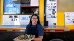 young hispanic woman st和ing in front of food truck with a tray of tacos